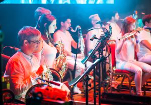 Side angle of musicians on stage, french horns, clarinet and bass clarinet, some musicians playing, lit in red and blue