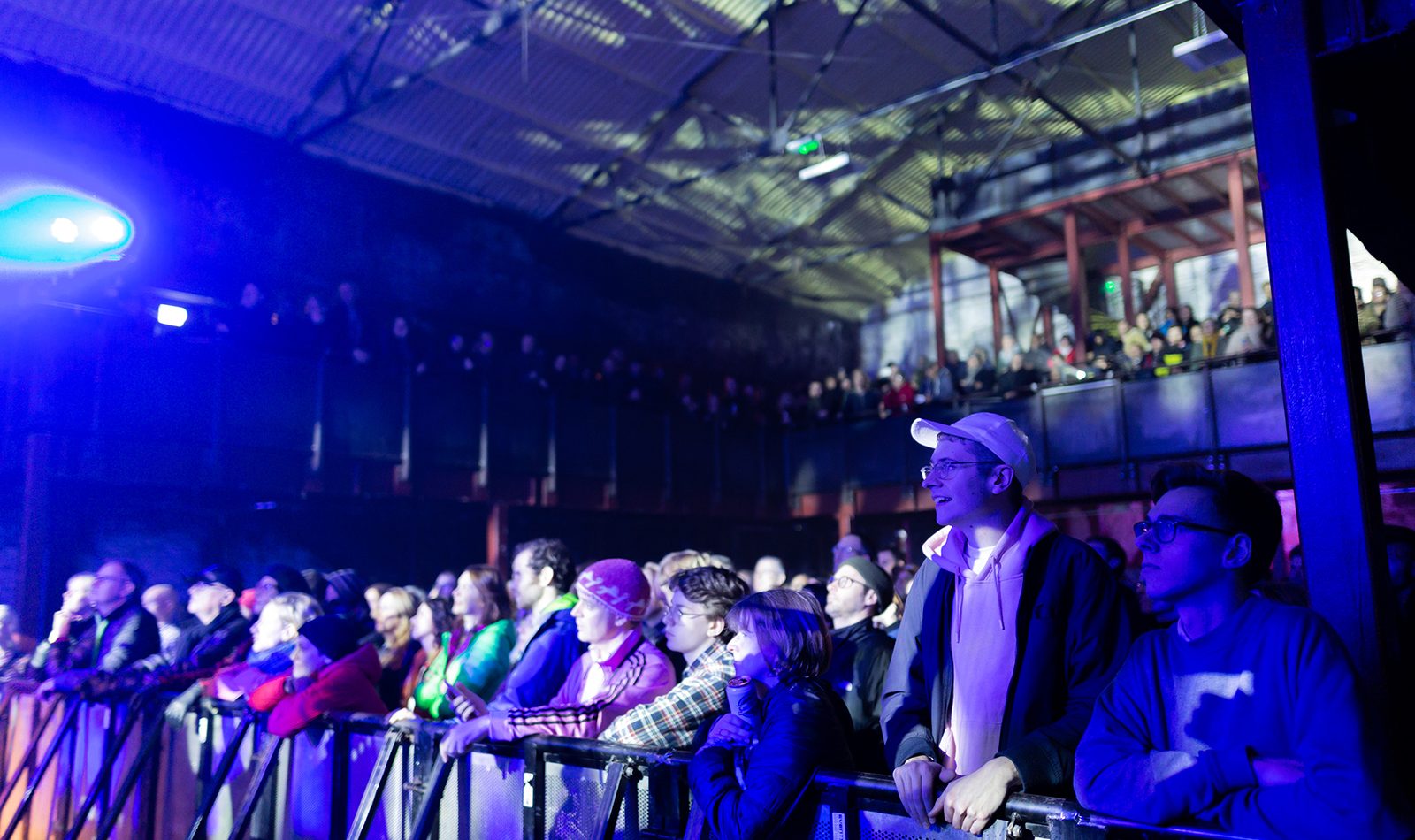 A gig crowd in a warehouse setting, dark, blue lighting, the standing audience watches an unseen stage