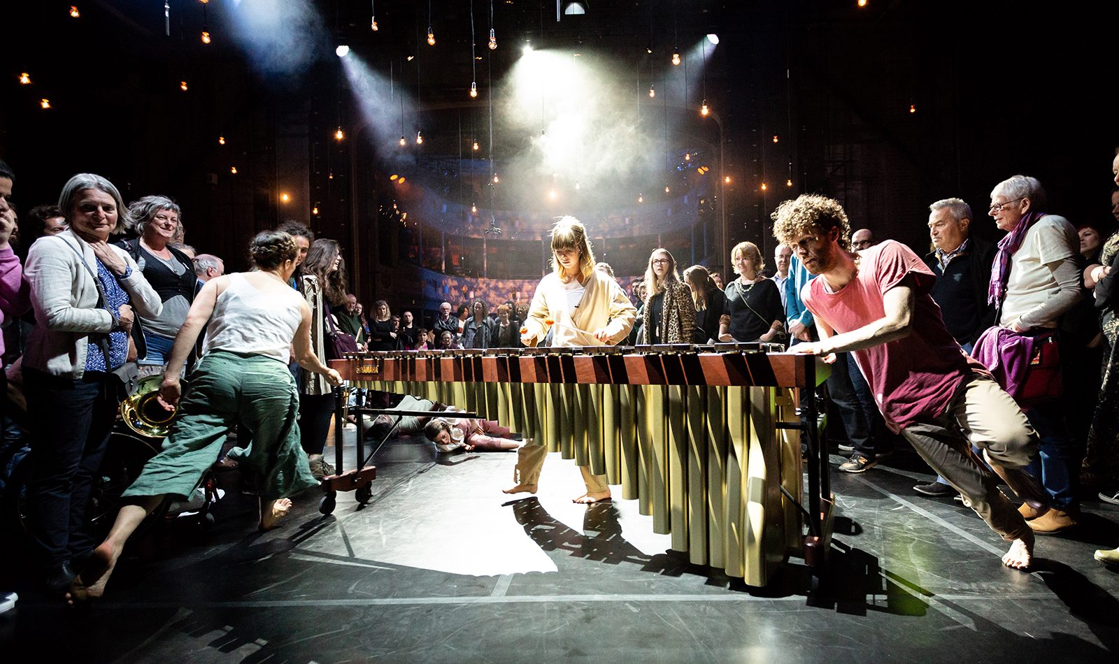 A female musician plays the marimba on stage, a male and a female dancer twirl it around from either end. Audience on stage look on.