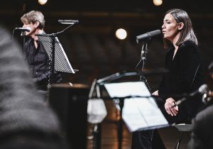 Brett Anderson, a light-skinned man in his fifties with short black-gray hair, and Nadine Shah, a light-skinned woman in her thirties with black shoulder-length hair, sit on a stage next to one another as they sing into microphones.