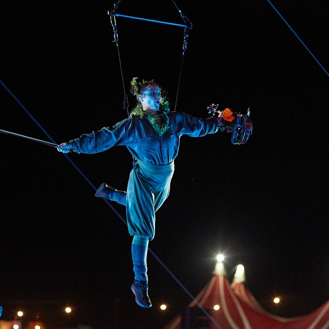A woman is suspended in mid-air by a harness attached aroudn her waist. She is wearing dramatic white-and-blue makeup and a blueovershirt with furry green trim, trousers, and shoes. In her left hand is a viola and in the other is a bow. Both of her arms are outstretched as she swings through the air. A white spotlight creates sharp relief against the night sky behind her. At the bottom of the image in the background the tips of a striped big-top style stretch tent are visible.