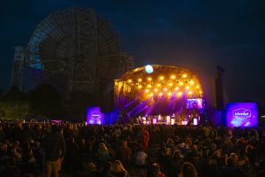 The main stage at Bluedot festival illuminated in purple and orange light. Visible behind the stage is the colossal Lovell Telescope, a huge metal structure with a dish telescope.