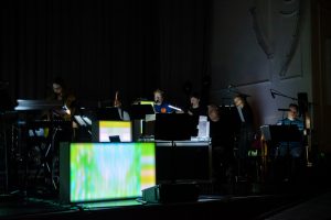 The wind section of Paraorchestra perform on stage in Edinburgh, a large green lightbox casting silhouettes across the wall behind them.