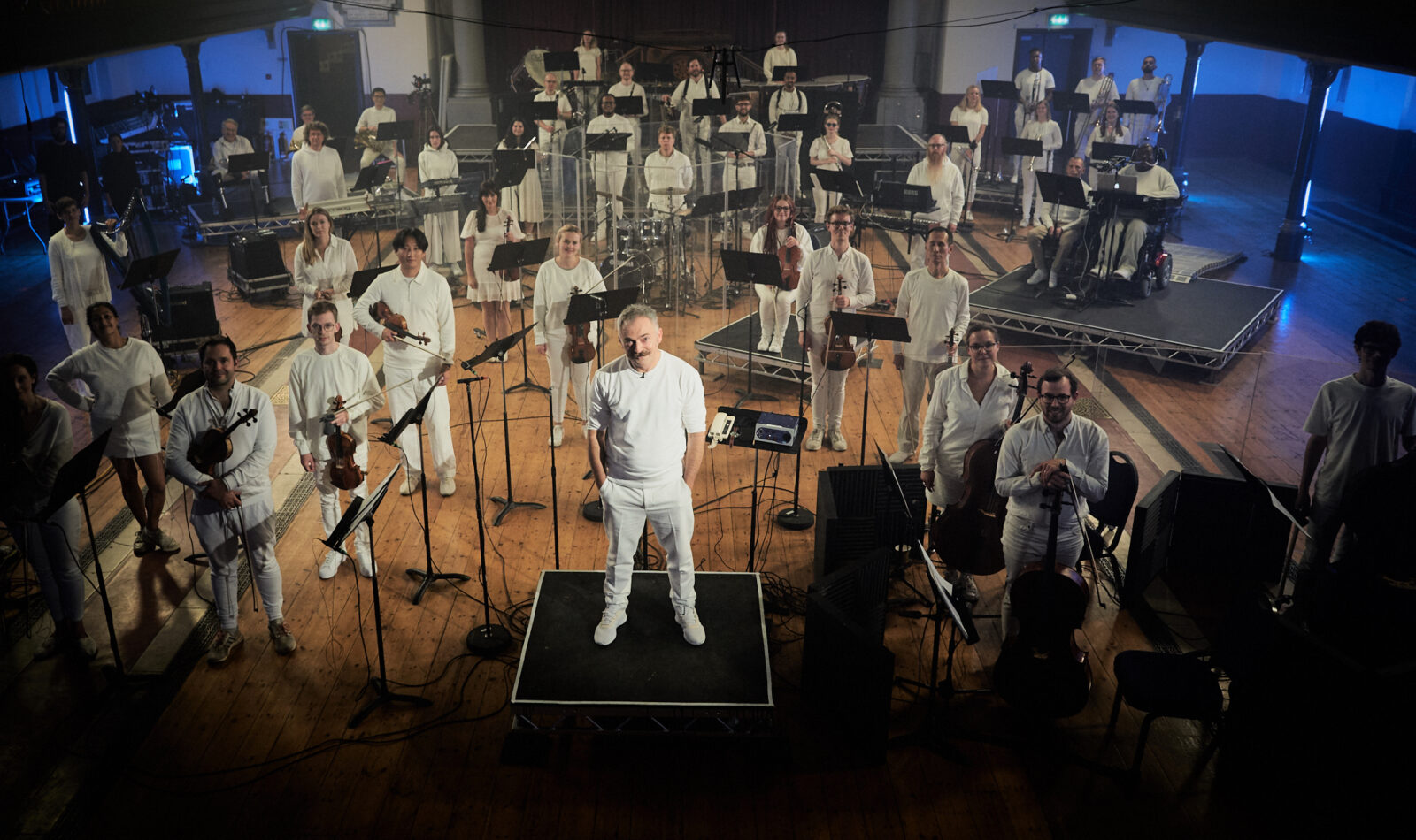 Charles Hazlewood, a man in his fifties with white-grey hair and moustache, stands confidently on a square riser in the centre of a performance hall, looking up confidently at the camera with one eyebrow slightly raised. Surrounding him are Paraorchestra musicians holding or sitting at their instruments and smiling. Both Charles and the musicans are dressed entirely in white.