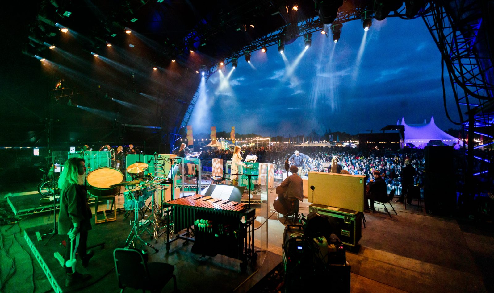 Taken from the back right of a festival stage, lighting in hues of green, within the orchestra onstage we can see a white female on percussion, the back of string players on cello and double bass. Centre stage a woman in a long white dress stands in front of a bank of synthesisers. Beyond the stage a crowd stands under a twilit cloudy sky, festival tents and lighting behind them.