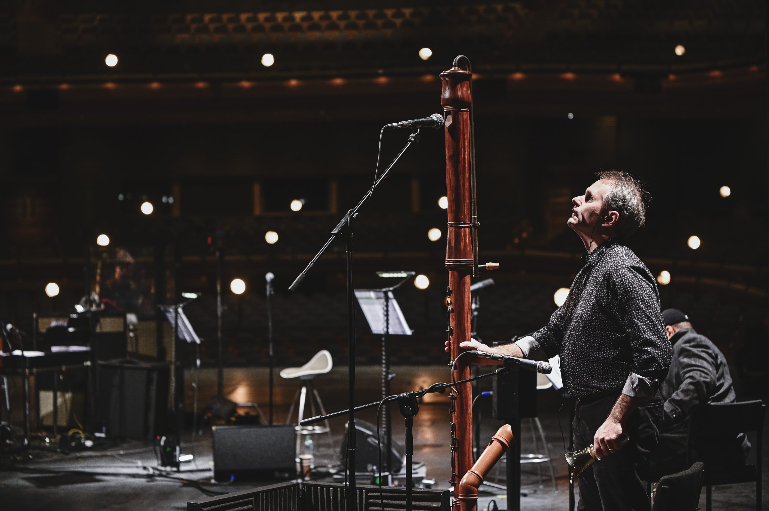 A tall woodwind instrument towers over a middle aged white male. He holds it with one hand and looks up to the mouthpiece at the top. A stage setting in the background shows music stands and microphones.
