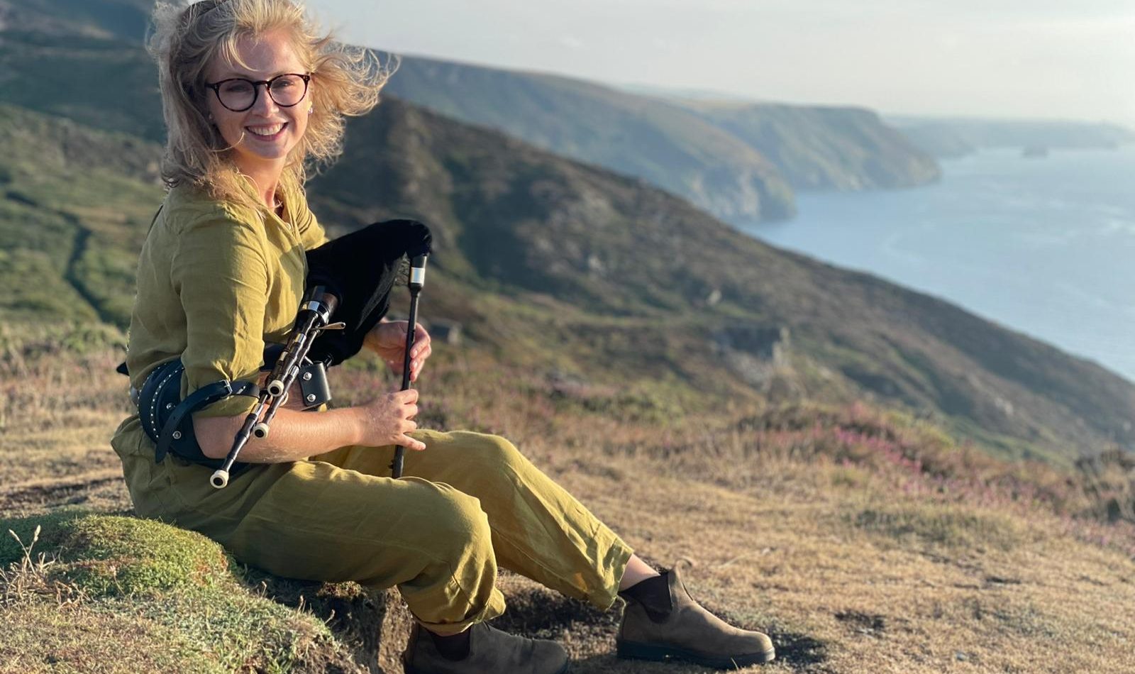 Hattie McCall Davies, a young white woman with blonde hair and glasses, sits on a clifftop with her scottish smallpipes. Behind her, a dramatic clifftop descends into the sea.