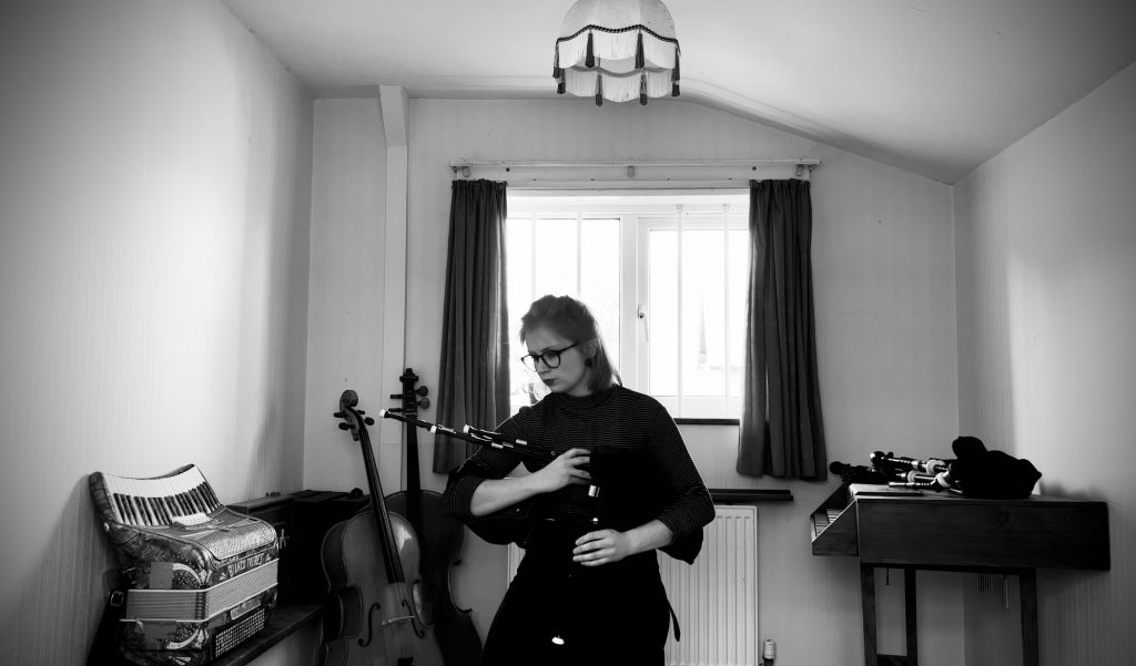 Hattie McCall Davies, a young white woman with blonde hair and glasses, plays the scottish smallpipes in a home studio. The room is lined with instruments, including an accordion, cello, and another set of pipes.