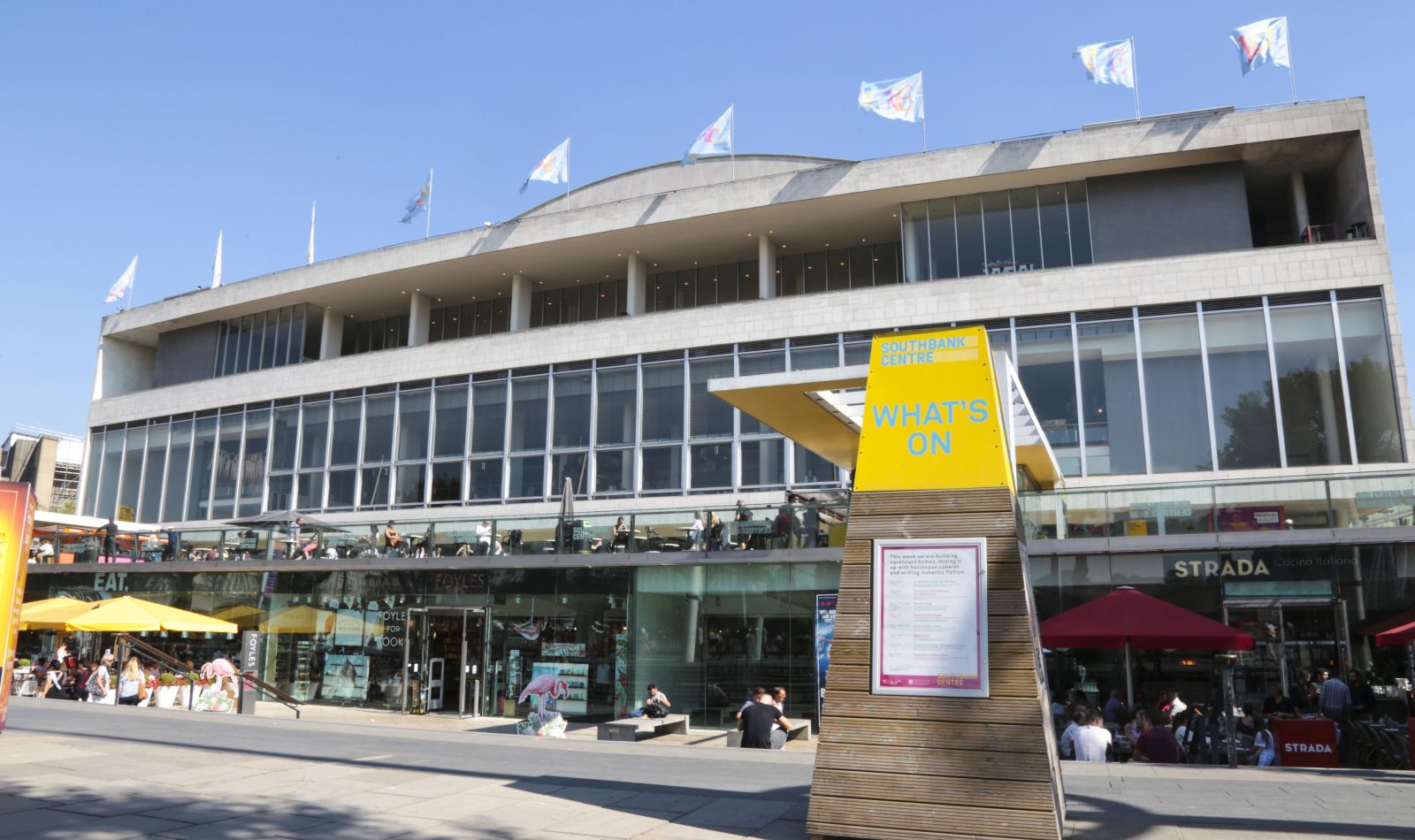 Photograph of Royal Festival Hall – a large concrete and glass building adorned with flags - beneath a cloudless blue sky