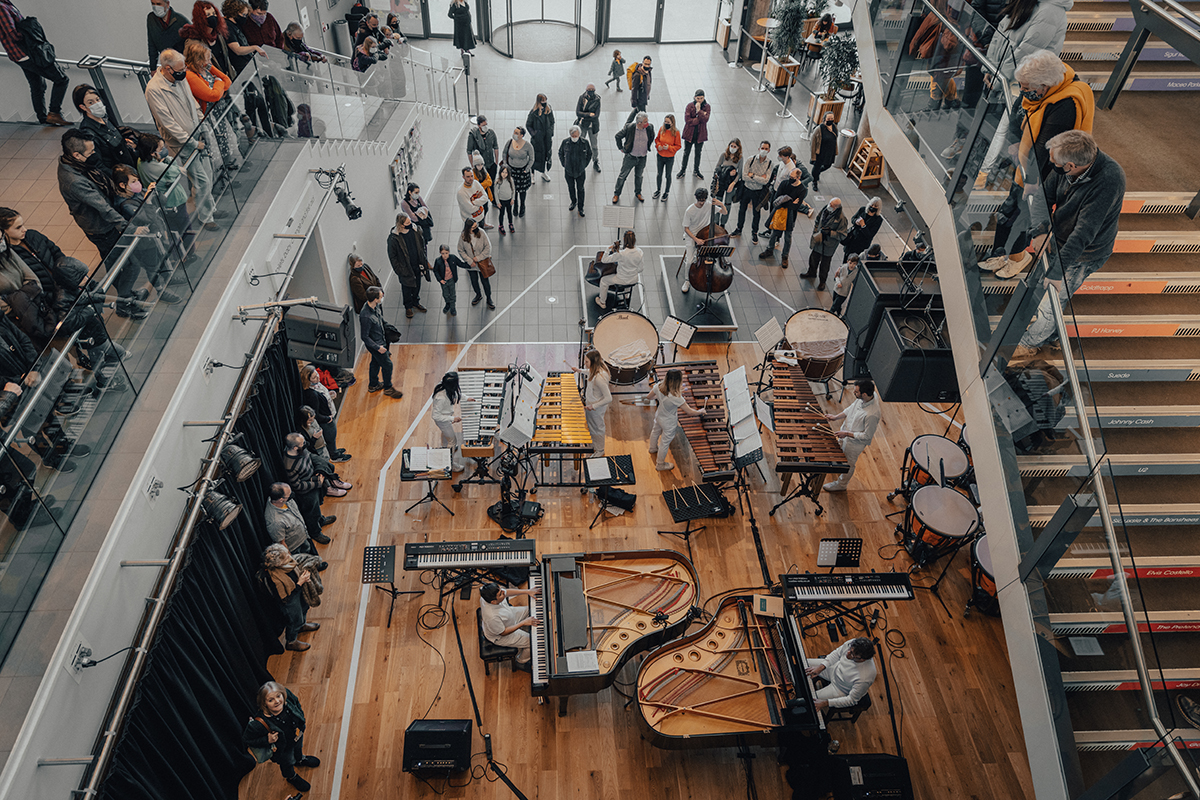 top-down view of pianists and a percussion section performing to a crowd in a bright atrium