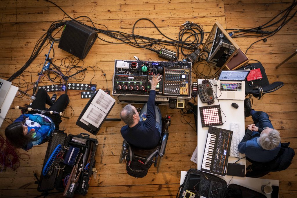 A birds-eye view of three musicians seated next to one another surrounded by wires, instruments, and equipment.